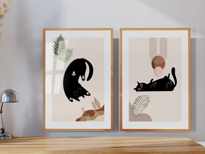 May include: Two framed prints with a minimalist design featuring black cats and abstract shapes in brown, pink, and beige tones. The prints have a light beige background and are framed in natural wood.