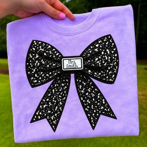 May include: A lavender sweatshirt featuring a large black bow with white speckles. The bow has a small rectangular tag in the center that reads "Mrs. Smith" in black text. The sweatshirt is a crew neck style.