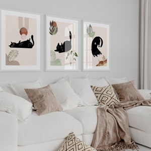 May include: Three framed prints with black cat illustrations on a white background. The prints are hung on a white wall above a white couch with brown throw pillows and a brown blanket.