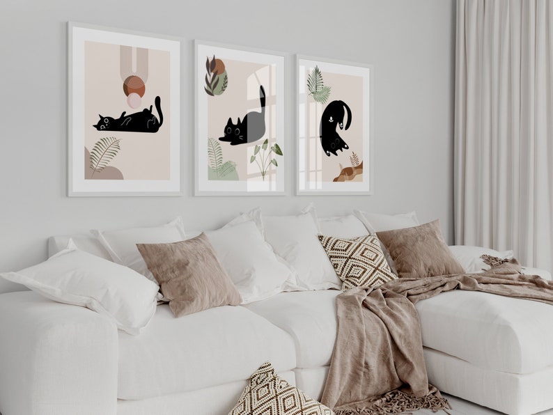 May include: Three framed prints with black cat illustrations on a white background. The prints are hung on a white wall above a white couch with brown throw pillows and a brown blanket.