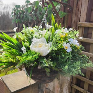 May include: A hanging basket filled with white peonies, small white and yellow flowers, and various green foliage. The basket is made of woven brown material and is suspended from a metal hook. The background includes a wooden fence and greenery.