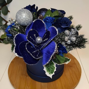 May include: A festive floral arrangement in a navy blue round box. The arrangement features deep blue velvet flowers, blue roses, silver glitter ornaments, and faux evergreen branches. The arrangement sits on a round wooden base.