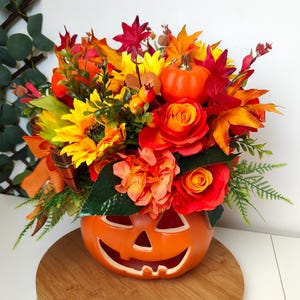 May include: A Halloween-themed floral arrangement in an orange pumpkin-shaped container. The display includes sunflowers, roses, and autumn leaves in vibrant red, orange, and yellow hues. A small pumpkin is also included in the arrangement.