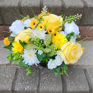 May include: A yellow and white artificial flower arrangement in a black pot. The arrangement features roses, daisies, and other small flowers.