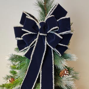 May include: A large navy blue velvet bow with silver trim, displayed on a Christmas tree. The bow's loops and tails are prominent, and the silver trim adds a festive sparkle. Pinecones are visible.