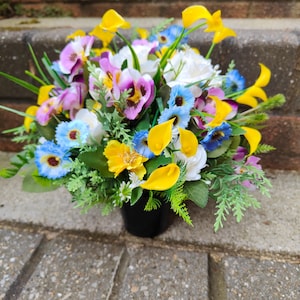 May include: A vibrant floral arrangement in a black pot. The bouquet features a mix of colorful artificial flowers, including yellow calla lilies, purple pansies, white roses, and blue daisies, with green foliage. The arrangement is set against a gray brick background.