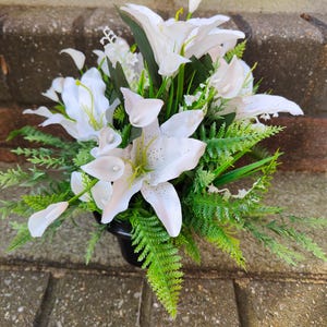 May include: A floral arrangement of white lilies and calla lilies, with green fern and foliage. The flowers are in a black container, creating a natural and elegant display.