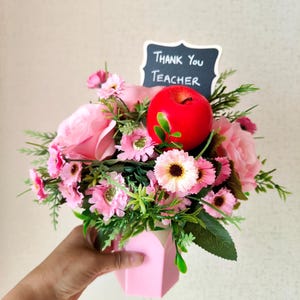 May include: A pink floral arrangement with a red apple and a chalkboard sign that reads "Thank You Teacher." The arrangement includes pink roses, daisies, and greenery, presented in a pink geometric vase.