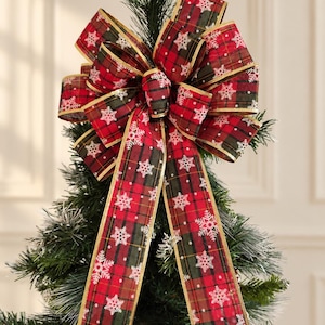 May include: A decorative Christmas tree topper bow featuring a red and dark green tartan pattern with white snowflake prints. The bow has multiple loops and two long hanging tails, all finished with a metallic gold wire edge trim.