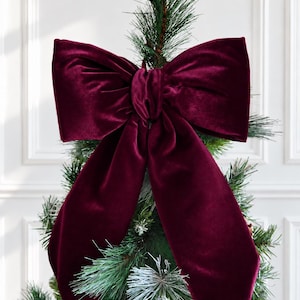 May include: A large, deep burgundy velvet bow with two long, pointed tails. The bow is tied in a classic style with a central knot and is positioned at the top of a green, artificial pine tree branch with frosted needle tips.