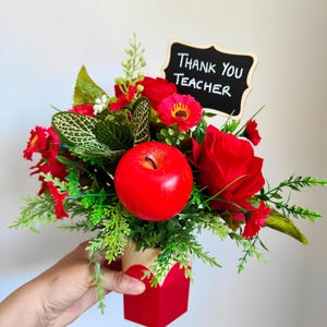 May include: A red and green floral arrangement in a red square vase, featuring a large red apple and a small chalkboard sign that reads "Thank You Teacher". The arrangement includes red roses and other red flowers.