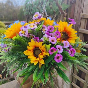 May include: A hanging basket overflowing with vibrant artificial flowers. The arrangement features large yellow sunflowers, purple and pink daisies, and blue and purple pansies, complemented by green foliage. The basket hangs from a metal hook and chain, creating a colorful display.