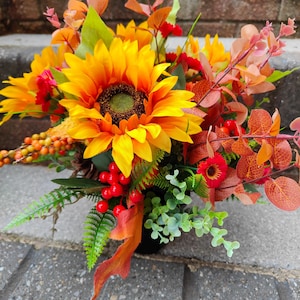 May include: A floral arrangement featuring a large yellow sunflower, smaller red flowers, orange berries, and various green and rust-colored foliage. The bouquet includes sprigs of eucalyptus, fern-like leaves, and red berries, creating a dense, multi-textured display of autumn-inspired colors and botanical elements.