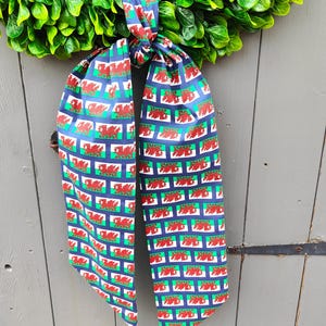 May include: A green wreath with a blue and red patterned ribbon. The ribbon features a repeating design of red dragons on a blue background, framed by green and white rectangles. The wreath is hanging on a gray wooden door.