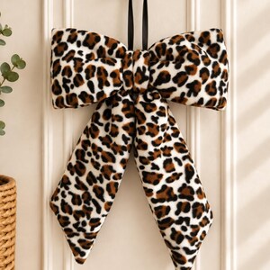 May include: A decorative bow made of soft, plush fabric featuring a brown, black, and white leopard print pattern. The bow is suspended by a thin black ribbon from a gold-coloured circular wall hook, with two long, pointed fabric tails hanging down.