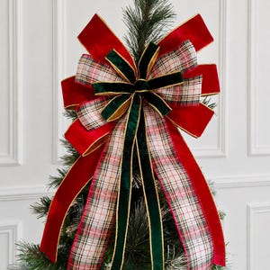 May include: A decorative Christmas tree topper bow featuring layered ribbons in solid red velvet, dark green velvet, and a cream-coloured plaid pattern with thin red and green lines. The bow has long, flowing tails and gold-trimmed edges.