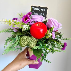 May include: A floral arrangement in a purple geometric vase, featuring a red apple, purple roses, and greenery. A small chalkboard sign reads "Thank You Teacher".