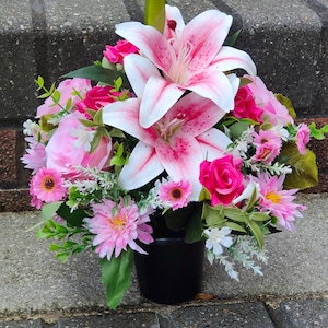 May include: A floral display in a black pot. The arrangement includes large pink and white lilies, pink roses, and other pink and white flowers with green leaves. The pot is placed on a grey brick surface.