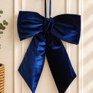 May include: A deep navy blue velvet bow ornament featuring a central knot and two long, wide tails hanging downwards. The bow is suspended from a matching navy blue ribbon loop, designed to be hung from a small, round, metallic gold wall hook.
