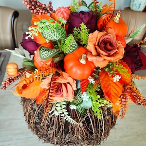 May include: A decorative arrangement featuring orange pumpkins with gold stems, deep burgundy and burnt orange fabric roses, green foliage, and brown berry sprigs, all set within a round, woven brown twig base for a seasonal display.
