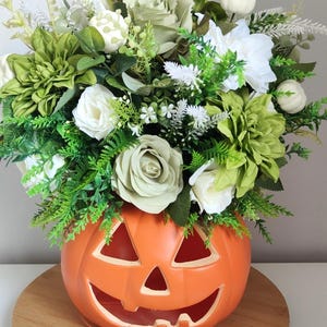 May include: A bright orange ceramic jack-o'-lantern pumpkin container filled with an arrangement of white and sage green artificial roses, dahlias, small white pumpkins, and various green foliage sprigs and ferns.
