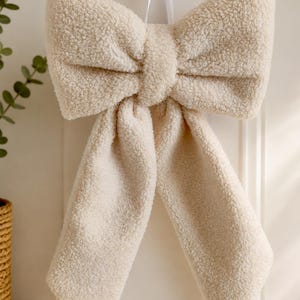 May include: A large, decorative bow made of cream-colored, textured sherpa fleece fabric. The bow features two wide, looped sections at the top and two long, hanging tails at the bottom, suspended by a thin white ribbon from a small gold hook.