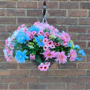 May include: A hanging basket filled with artificial flowers in pink, blue, and white. The woven basket hangs from a metal hook and chain against a brick wall. The flowers include dahlias and other varieties.
