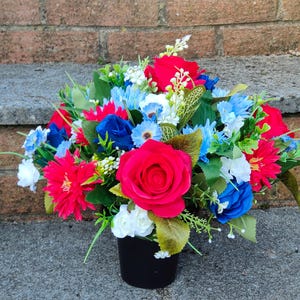 May include: A red, white, and blue artificial flower arrangement in a black pot. The arrangement features roses, daisies, and other flowers.