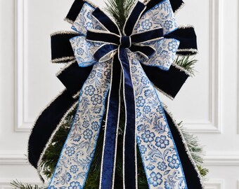 May include: A decorative tree topper bow featuring a combination of solid navy blue velvet ribbon and white ribbon with a blue floral pattern. The edges of the ribbons are trimmed with silver metallic tinsel, and the bow has long cascading tails.