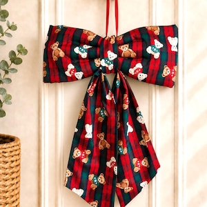May include: A large decorative fabric bow featuring a red and dark green tartan pattern with a repeating print of small brown and white teddy bears wearing jumpers. The bow is suspended by a thin red ribbon from a round gold-coloured wall hook.