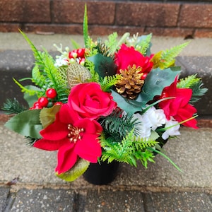 May include: A festive floral arrangement in a black pot. The arrangement features vibrant red poinsettias, roses, and berries, complemented by green foliage and a golden pinecone. The flowers are set against a backdrop of brick and concrete.