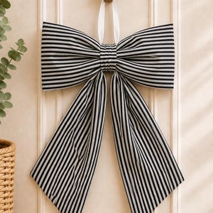 May include: A large decorative bow featuring a pattern of thin black and white vertical stripes. The bow is constructed with two wide horizontal loops and two long, pointed hanging tails, suspended from a small white ribbon loop at the center.