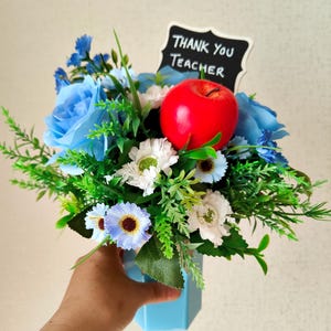 May include: A floral arrangement in a light blue vase, featuring blue roses, white and blue daisies, and green foliage. A red apple and a small chalkboard sign that reads "THANK YOU TEACHER" are included.