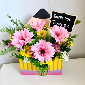 May include: A floral arrangement in a yellow box with pink and purple stripes, filled with pink gerbera daisies, yellow flowers, and greenery. A small chalkboard sign reads "Thank You Teacher."