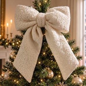 May include: A large, cream-coloured bow made of a textured, sherpa-like fabric. The bow is positioned at the top of a green artificial Christmas tree, which is decorated with warm white string lights and several round, metallic gold ornaments.
