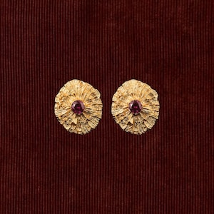 May include: A pair of circular gold earrings with a textured, radiating fan-like surface pattern. Each earring features a single, round, deep red gemstone set securely in the center of the textured gold metal disc design.