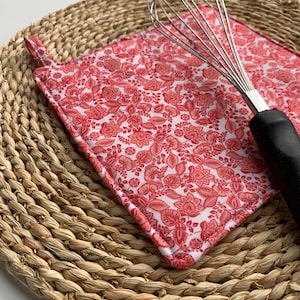May include: Red and white floral patterned pot holder with a woven straw placemat and a silver whisk.