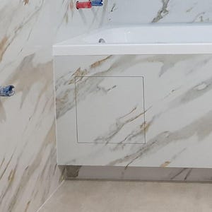 Könnte beinhalten: Badezimmerinterieur mit weißer Badewanne und Wandfliesen mit Marmormuster. Die Wand hat eine quadratische Zugangsklappe mit schwarzer Umrandung. Sanitärrohre mit roten und blauen Enden sind sichtbar.