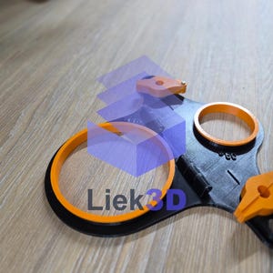 Op de afbeelding: Een zwart en oranje 3D-geprint object met de tekst "Liek3D". Het object heeft twee oranje ringen en twee oranje klemmen. Het object ligt op een houten oppervlak.