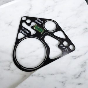 Peut inclure: Outil en métal noir avec diverses découpes circulaires et angulaires. Il comporte un niveau intégré et des mesures en millimètres, dont 20 mm, 50 mm, 60 mm, 110 mm et 120 mm. L'outil est conçu pour la précision et la mesure.