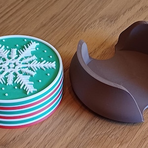 May include: A stack of round coasters featuring a green top with a white snowflake design and alternating red, white, and green striped sides, accompanied by a dark brown circular holder with a curved cutout design for storage.