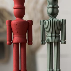 Puede incluir: Dos figuras de soldados de pie sobre una base plana. La figura de la izquierda es de color rojo sólido con una superficie texturizada y detalles de uniforme. La figura de la derecha es verde salvia y está hecha de listones verticales.