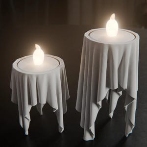 May include: Two light grey candle holders with a draped fabric appearance, each featuring a circular top and a glowing white LED flame insert. The structures have a smooth, matte finish with deep folds that mimic hanging cloth.