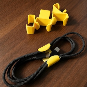 Könnte beinhalten: Ein schwarzes geflochtenes Ladekabel mit Metallanschlüssen, das von zwei gelben Kunststoff-Kabelclips gehalten wird. Drei weitere gelbe Kunststoff-Kabelclips sind in einer Gruppe in der Nähe angeordnet und haben jeweils ein U-förmiges Design.