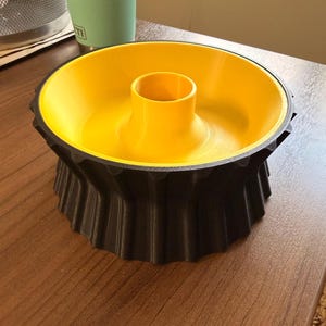 Peut inclure: Un objet circulaire en forme de bol avec un intérieur jaune vif et un pilier cylindrique central. L'extérieur présente un design noir, verticalement nervuré ou cannelé, qui s'évase vers le bord supérieur, créant un contraste marqué.