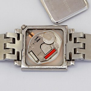 May include: A silver-toned wristwatch with a square face, revealing the internal mechanism. The watch band is also silver and has a unique, geometric design. The watch's back cover is detached, showing the battery and other components.