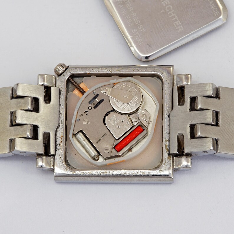 May include: A silver-toned wristwatch with a square face, revealing the internal mechanism. The watch band is also silver and has a unique, geometric design. The watch's back cover is detached, showing the battery and other components.
