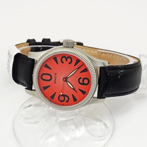 May include: A silver Raketa watch with a red face and black numerals. The watch has a black leather strap and a textured bezel. The watch face displays the word "Raketa".