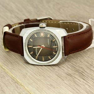 Könnte beinhalten: Eine silberfarbene Vintage-Armbanduhr mit braunem Lederarmband. Das Zifferblatt der Uhr ist dunkel mit goldfarbenen Markierungen und Zeigern, einschließlich eines roten Sekundenzeigers. Die Uhr zeigt das Datum und die Marke "Wostok".
