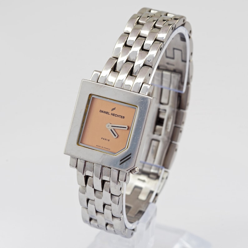 May include: A silver-toned Daniel Hechter watch with a square face and a linked bracelet. The watch face is peach-colored with black hands and the text "DANIEL HECHTER PARIS". The watch is made in France.